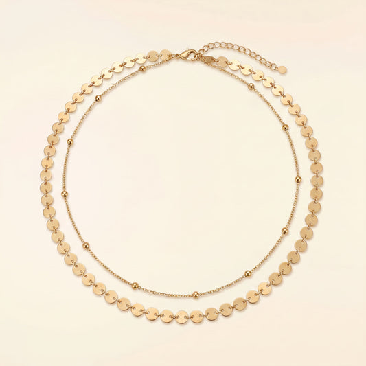 A gold-toned double-strand necklace featuring a layered design with flat circular disc links on the outer layer and smaller bead links on the inner layer, displayed on a light cream background.