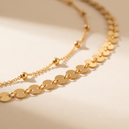 A close-up of a gold-toned necklace with flat circular disc links and a few small bead accents, photographed on a warm off-white background with soft shadows.