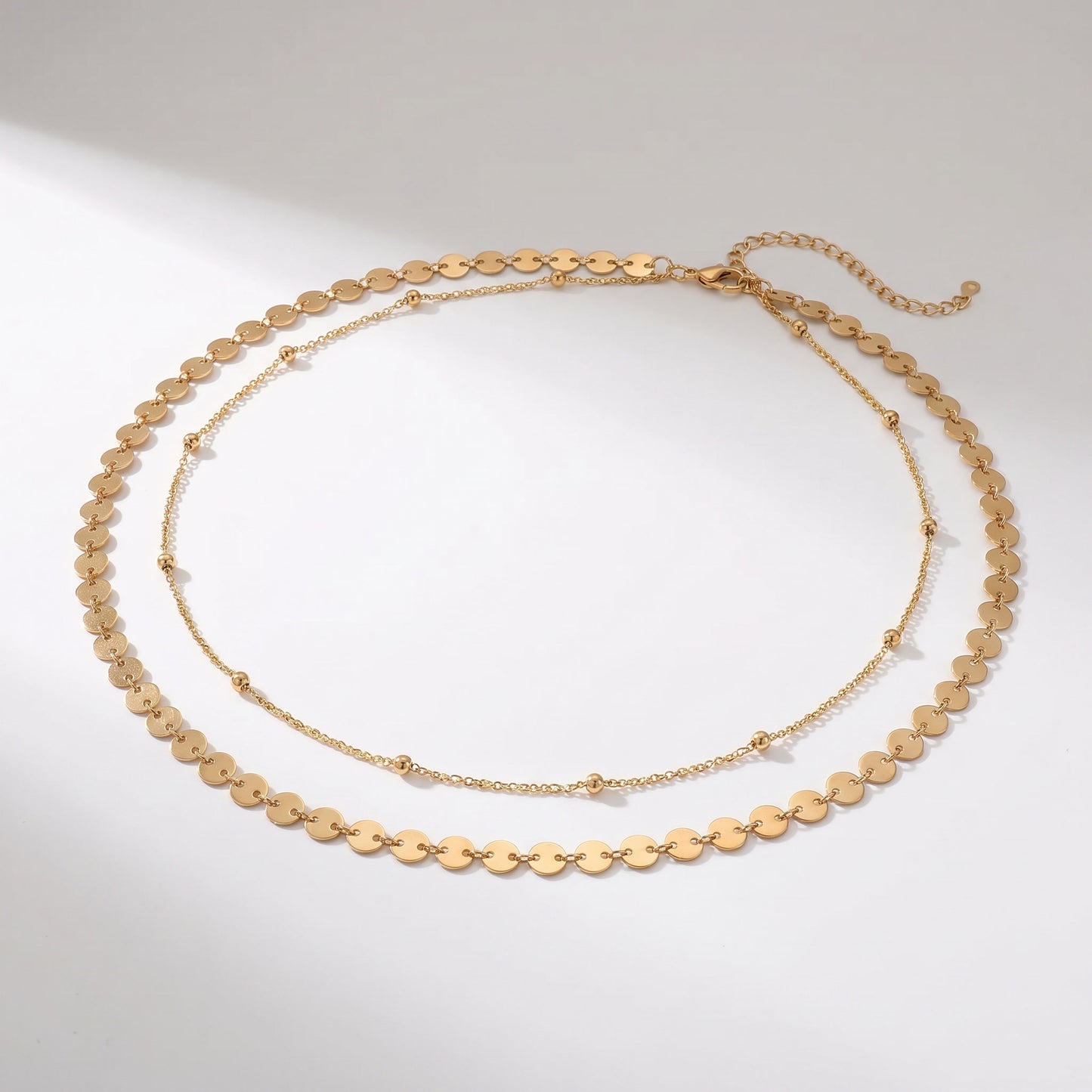 A gold-toned layered necklace featuring a short chain of flat oval disc links and a longer delicate chain with small bead accents, displayed on a light gray background.