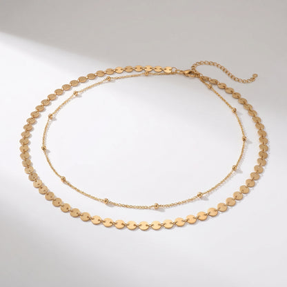 A gold-toned layered necklace featuring a short chain of flat oval disc links and a longer delicate chain with small bead accents, displayed on a light gray background.