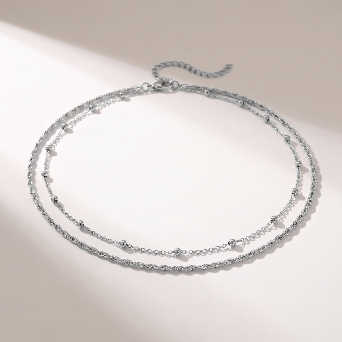 A delicate double-layered silver necklace with a textured twisted chain on the top layer and a fine beaded chain on the bottom layer, displayed on a warm off-white background with soft diagonal shadows.