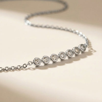A silver chain necklace with seven bezel-set round clear stones, photographed on a warm beige background.