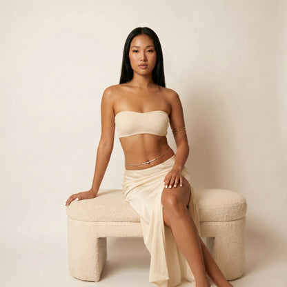 A woman poses on a textured cream-colored bench wearing a matching cream-colored two-piece set consisting of a strapless bandeau top and a draped high-slit skirt, accessorized with delicate silver chain bracelets.