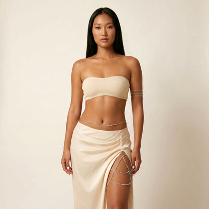 A woman modeling a cream-colored ribbed tube top and matching draped maxi skirt with silver chain accents, set against a plain beige studio background.