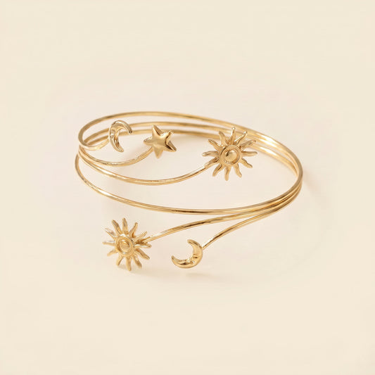 A delicate gold wrap bangle bracelet featuring celestial motifs including suns, moons, and a star, displayed on a warm cream background.