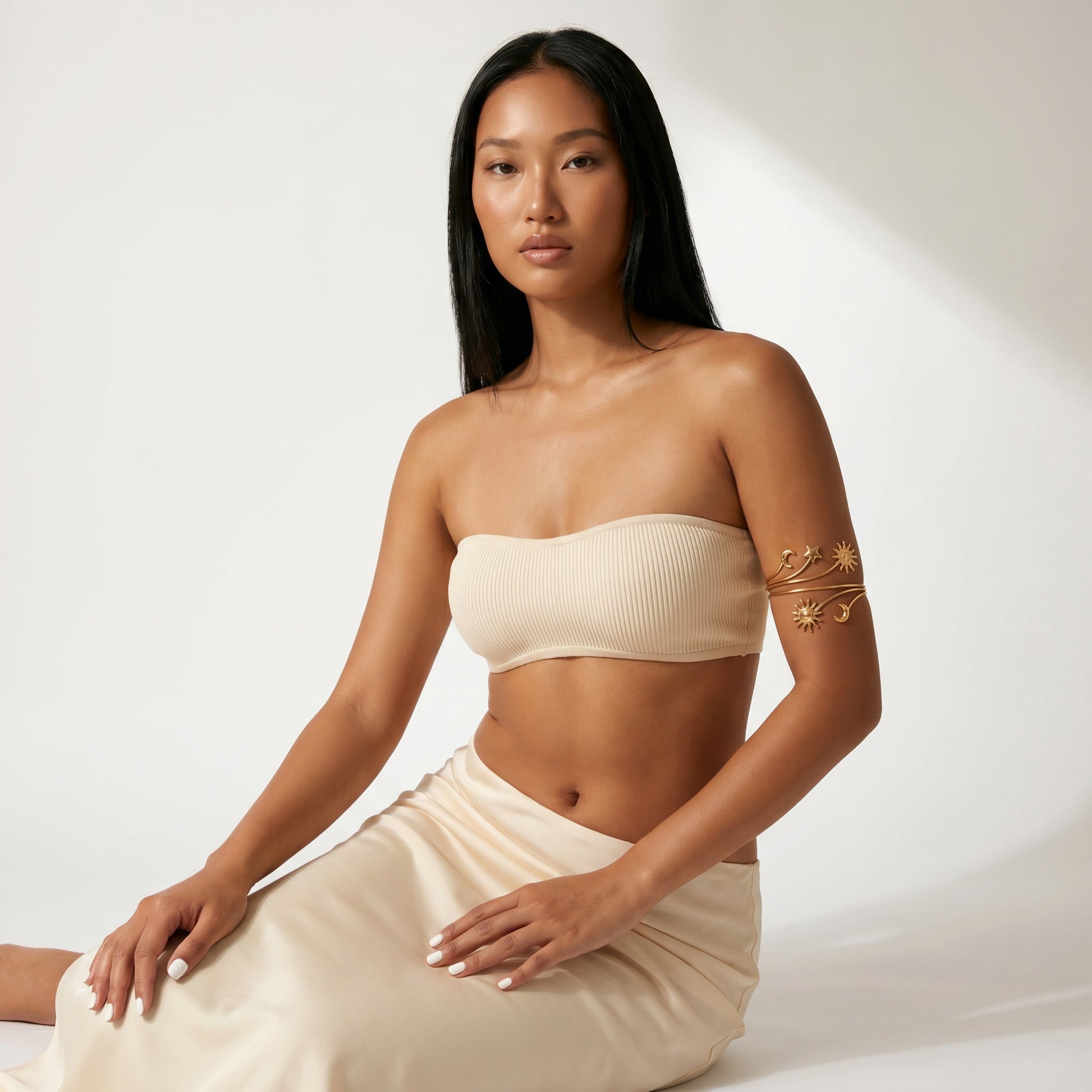 A woman wearing a beige ribbed strapless bandeau top and matching beige skirt, seated against a white studio background with soft diagonal shadows. She has a gold leaf-patterned armband accessory on her upper arm.