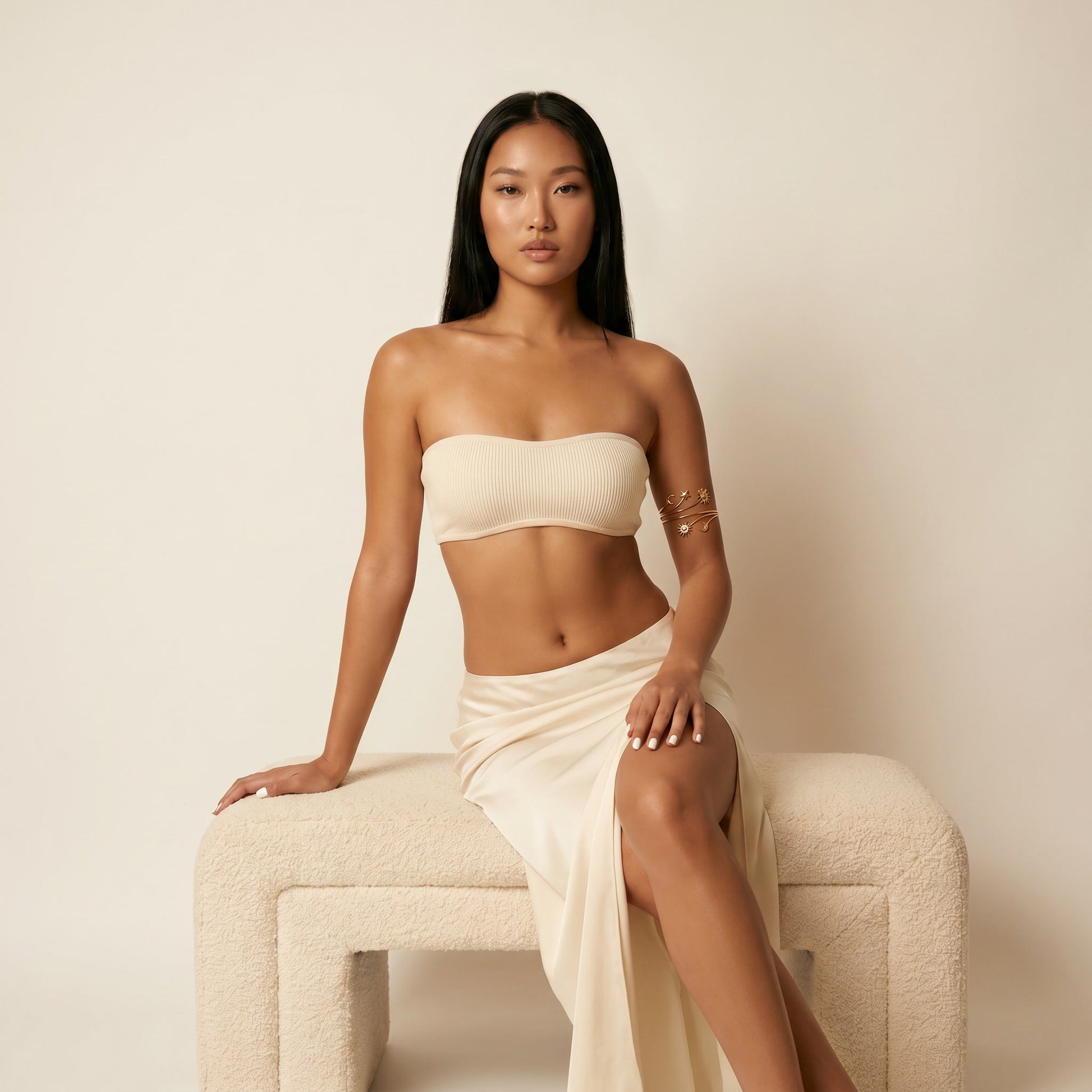 A woman with long black hair sits on a textured cream-colored bench against a solid cream background, wearing a matching beige ribbed bandeau top and draped maxi skirt.