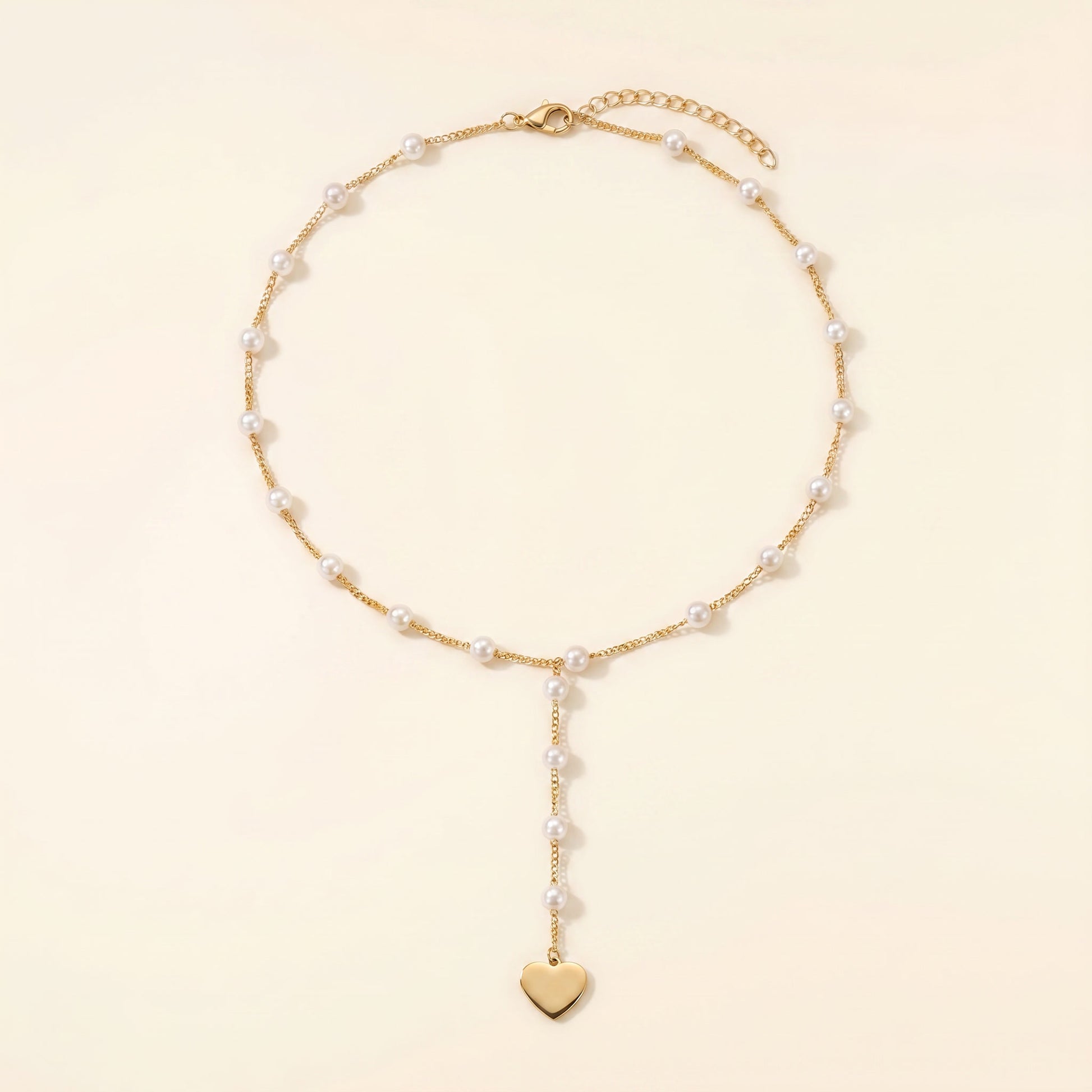 A delicate gold chain necklace featuring small white pearls spaced along the chain and a dangling gold heart charm at the bottom, displayed on a warm cream background.