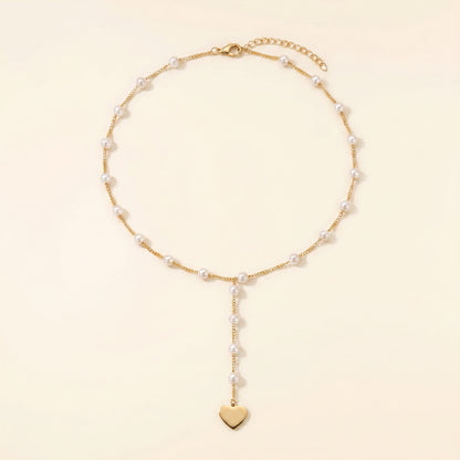 A delicate gold chain necklace featuring small white pearls spaced along the chain and a dangling gold heart charm at the bottom, displayed on a warm cream background.