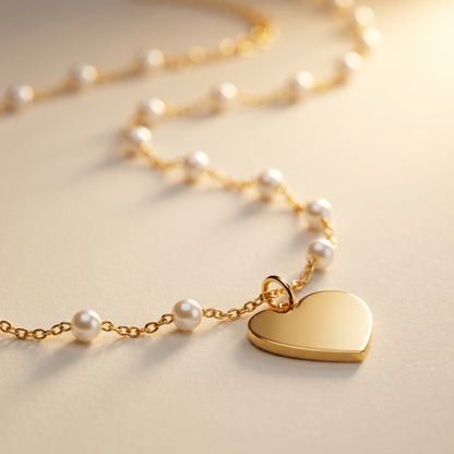 A gold heart pendant necklace with a delicate gold chain and small white pearl beads, displayed on a warm beige surface.