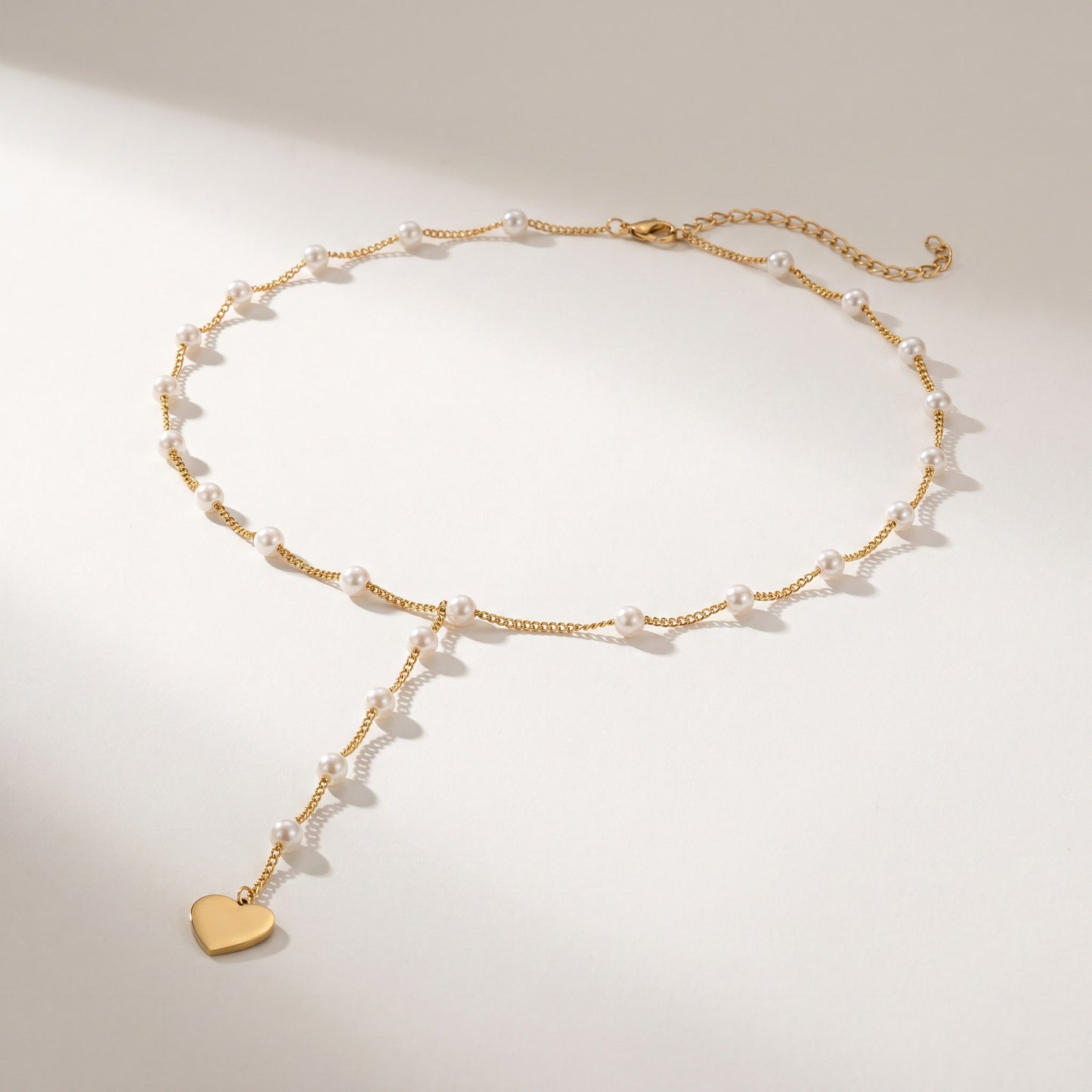 A delicate gold chain necklace with small white pearl beads and a dangling gold heart charm, displayed on a clean light beige background.