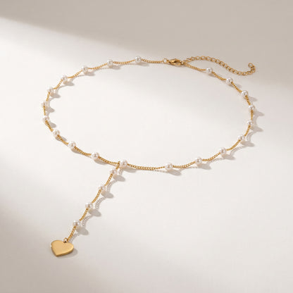 A delicate gold chain necklace with small white pearl beads and a dangling gold heart charm, displayed on a clean light beige background.