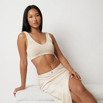 A woman models a matching two-piece knit and satin set in a warm cream or off-white color, consisting of a textured crop top and a draped high-slit skirt, accessorized with a delicate gold chain belt.