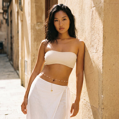 A woman stands in a sunlit stone alley wearing a white ribbed bandeau-style top and a white wrap skirt, with a delicate gold chain belt at her waist.