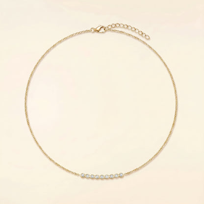 A delicate gold chain necklace with a small curved bar of clear round stones at the center, displayed on a light cream background.