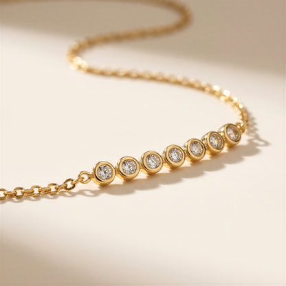 A gold chain necklace featuring seven bezel-set round clear gemstones on a warm cream background.