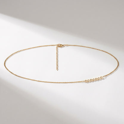 A delicate gold chain necklace with a small cluster of clear faceted beads on one side, displayed on a clean white surface with soft diagonal shadows.