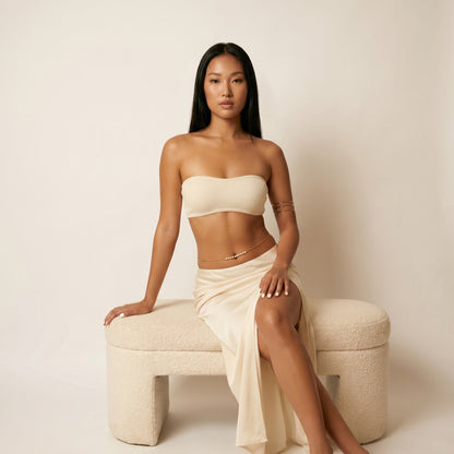 A woman poses on a textured cream-colored bench wearing a matching cream-colored two-piece set consisting of a strapless bandeau top and a draped high-slit skirt.