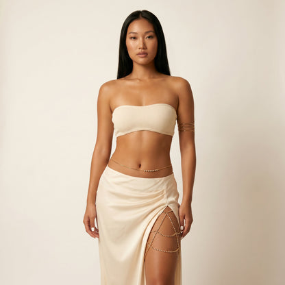 A woman modeling a cream-colored ribbed bandeau crop top and matching draped wrap skirt with gold chain accents, set against a plain beige studio background.