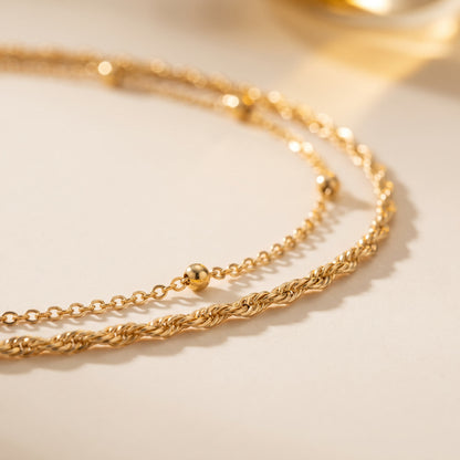 A double-strand gold chain necklace with small bead accents, displayed on a light cream background.