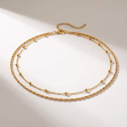 A delicate double-layered gold necklace with a textured twisted chain on the top layer and a fine beaded chain on the bottom layer, displayed on a warm off-white background with soft diagonal shadows.