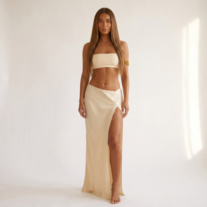 A woman stands barefoot in a studio wearing a matching beige two-piece set consisting of a strapless bandeau top and a high-slit maxi skirt, accessorized with gold chain details and bracelets.