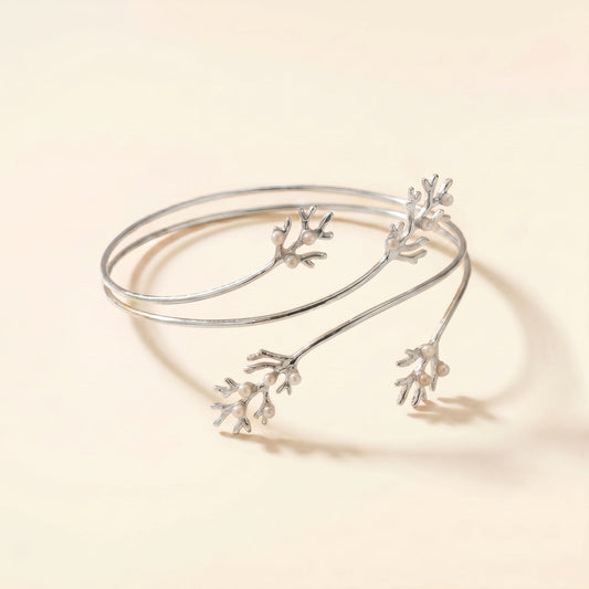 A delicate silver open cuff bangle featuring three small branching coral or branch-like motifs, photographed on a warm cream background.