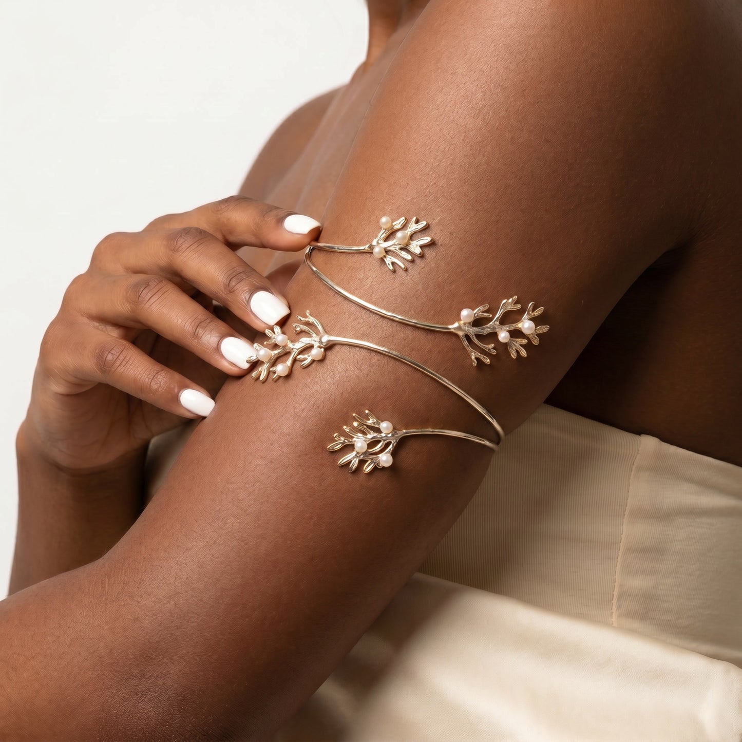 A close-up of a model's upper arm and hand wearing delicate gold-toned branch-shaped cuff bracelets with small white floral accents, against a soft light background.