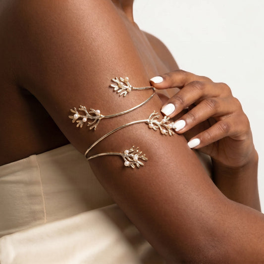 A close-up of a model's upper arm and hand wearing delicate gold-toned branch-shaped cuff bracelets with small white floral accents, against a soft light background.