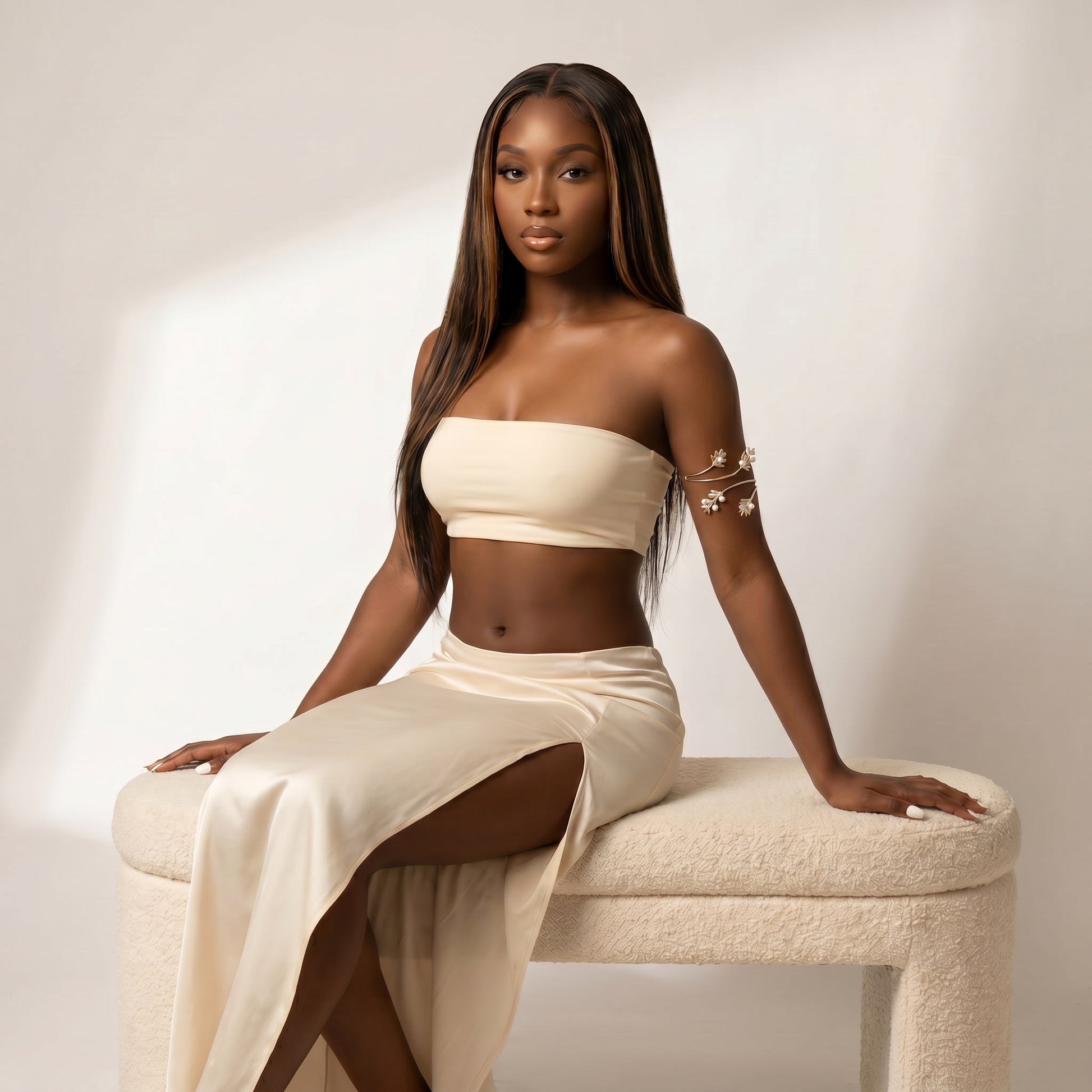 A woman with long dark hair sits on a cream upholstered bench wearing a matching cream two-piece outfit with a strapless bandeau top and a high-slit skirt. She has delicate gold floral arm bracelets on one arm, posed against a warm beige studio wall with soft diagonal shadows.