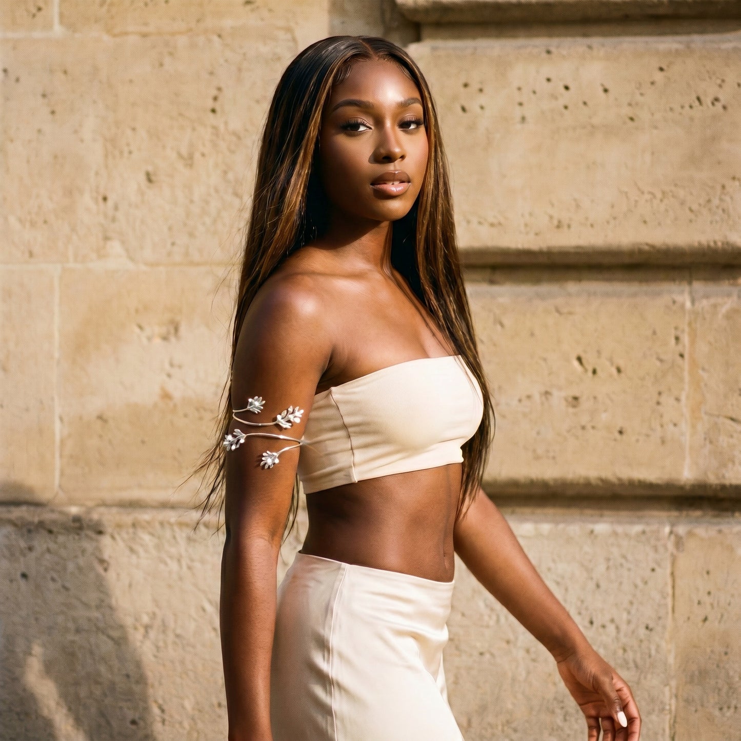 A woman posing outdoors against a beige stone wall, wearing a cream-colored two-piece set consisting of a strapless bandeau top with white floral embroidery on the shoulder and a matching high-waisted skirt.