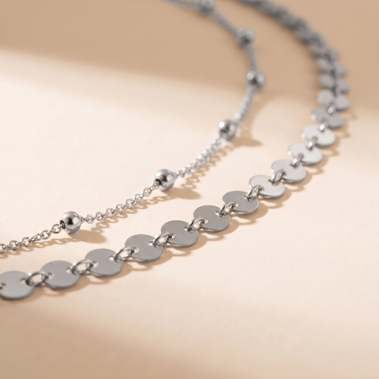 A close-up of a silver-toned necklace with flat circular disc links and a few small bead accents, photographed on a warm off-white background with soft shadows.