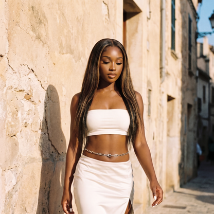 A woman with long dark hair walks down a sunlit stone alley wearing a white strapless tube top and a matching white wrap skirt with a small silver chain detail at the waist.