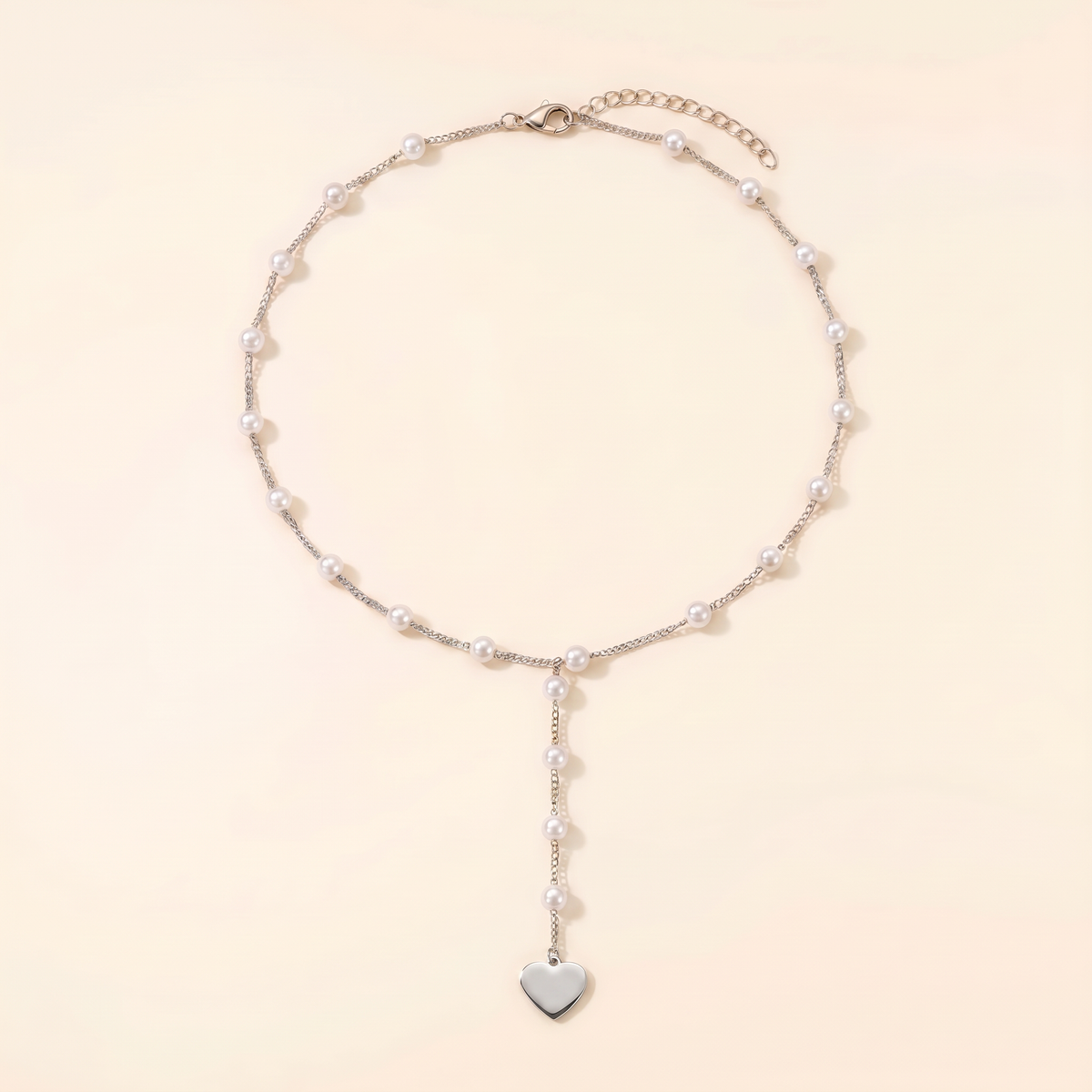 A delicate silver chain necklace featuring small white pearls spaced along the chain and a dangling silver heart charm at the bottom, displayed on a warm cream background.