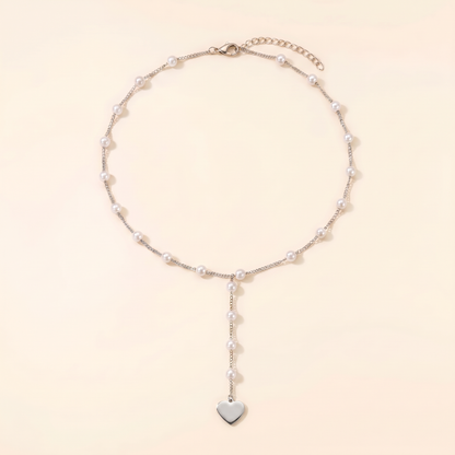 A delicate silver chain necklace featuring small white pearls spaced along the chain and a dangling silver heart charm at the bottom, displayed on a warm cream background.