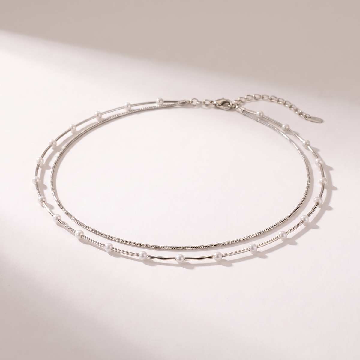 A delicate double-layered silver chain necklace with small white pearl accents, displayed on a clean, light beige background with soft shadows.