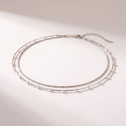 A delicate double-layered silver chain necklace with small white pearl accents, displayed on a clean, light beige background with soft shadows.