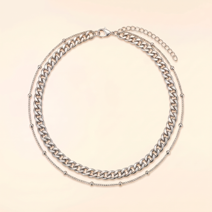 A silver-toned double-layer necklace featuring a chunky curb chain and a delicate beaded chain, displayed on a light cream background.