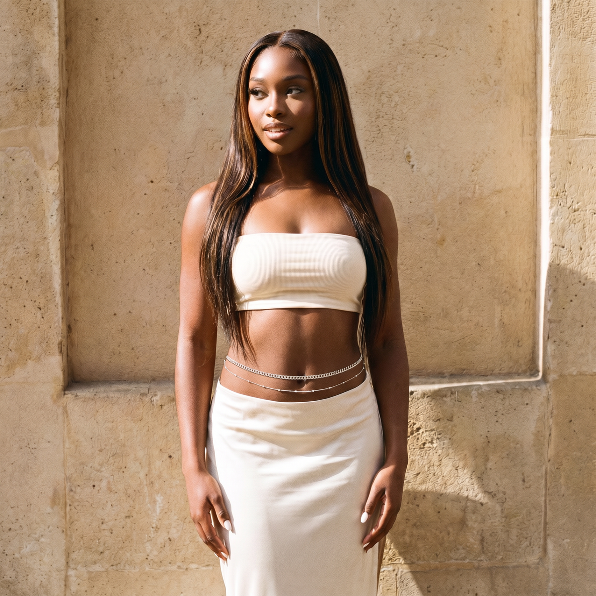 A woman with long dark hair stands against a textured beige stone wall, wearing a matching off-white two-piece outfit consisting of a strapless bandeau crop top and a high-waisted skirt, accessorized with a silver chain belt.