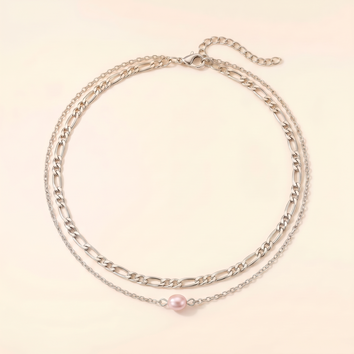 A double-layered silver chain necklace featuring a classic Figaro chain and a delicate fine chain with a single pink pearl pendant, displayed on a light cream background.