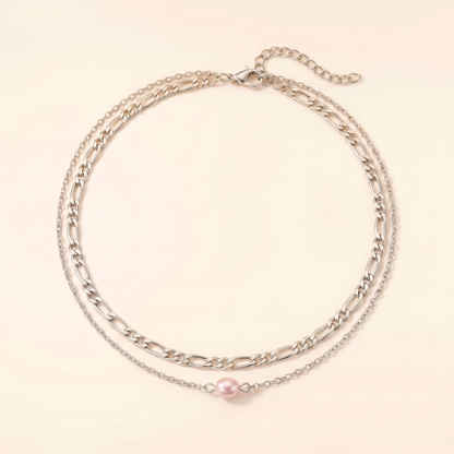 A double-layered silver chain necklace featuring a classic Figaro chain and a delicate fine chain with a single pink pearl pendant, displayed on a light cream background.