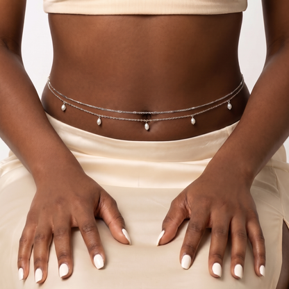 A close-up of a model's midsection wearing a delicate double-strand silver chain belly chain decorated with small white pearl-like beads, paired with a cream-colored satin skirt.