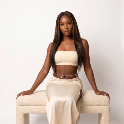 A woman with long, straight dark hair sits on a cream boucle bench against a plain white studio background. She is wearing a matching cream two-piece set consisting of a strapless crop top and a high-slit maxi skirt, accessorized with a thin silver chain belt.