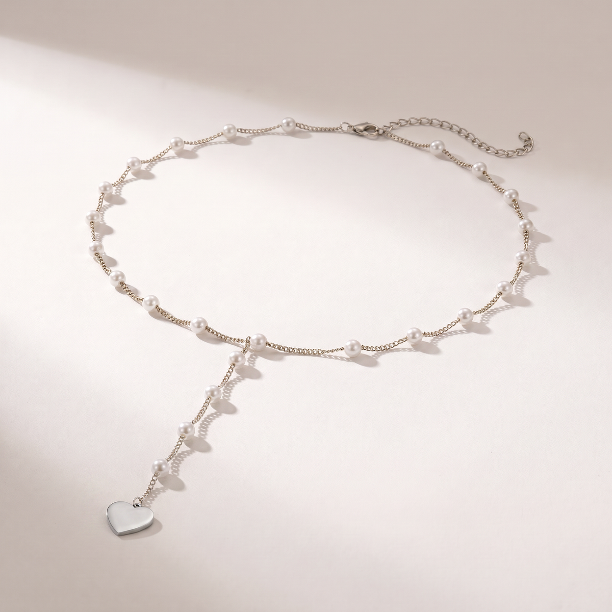 A delicate silver chain necklace with small white pearl beads and a dangling silver heart charm, displayed on a clean light beige background.