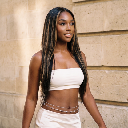 A woman with long, straight black hair with blonde highlights is wearing a white strapless tube top and matching white bottoms with a silver chain belt, standing against a beige stone wall.