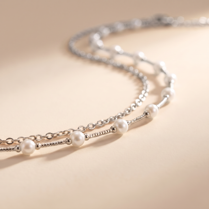 A delicate silver chain necklace with evenly spaced white pearl beads, photographed on a warm beige background with soft shadows.
