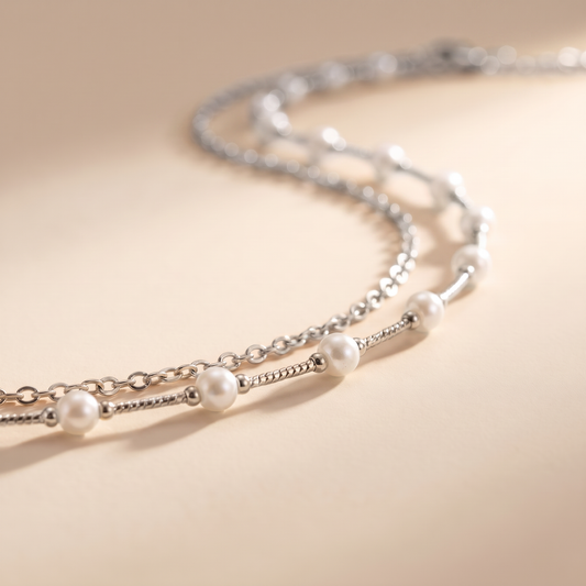 A delicate silver chain necklace with evenly spaced white pearl beads, photographed on a warm beige background with soft shadows.