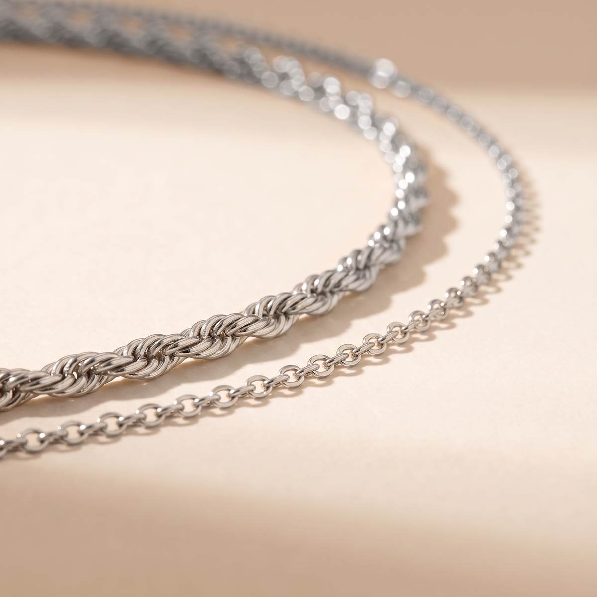 A close-up of a silver chain necklace draped in two parallel loops on a warm beige surface with soft shadows.
