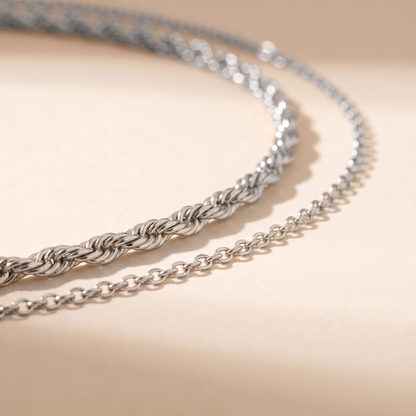 A close-up of a silver chain necklace draped in two parallel loops on a warm beige surface with soft shadows.
