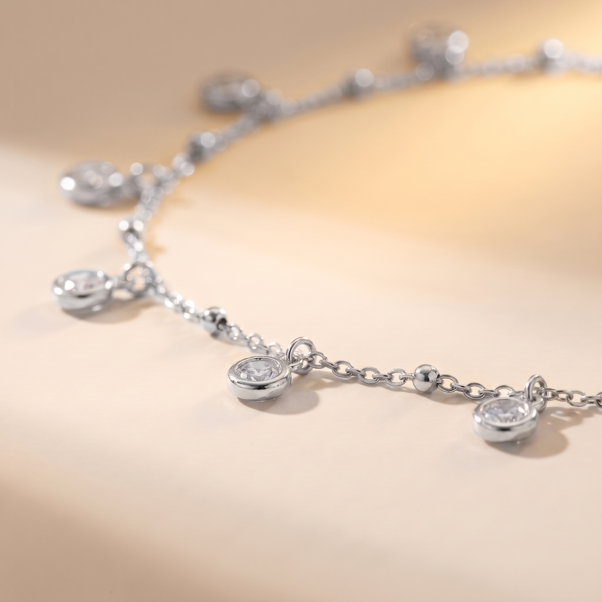 A close-up of a delicate silver chain necklace with small oval bezel-set clear stones, photographed on a warm cream background.